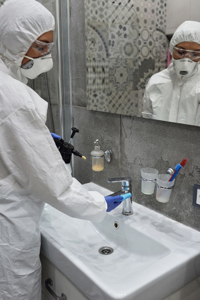 why-choose-us Person in protective gear disinfecting bathroom sink with spray bottle.