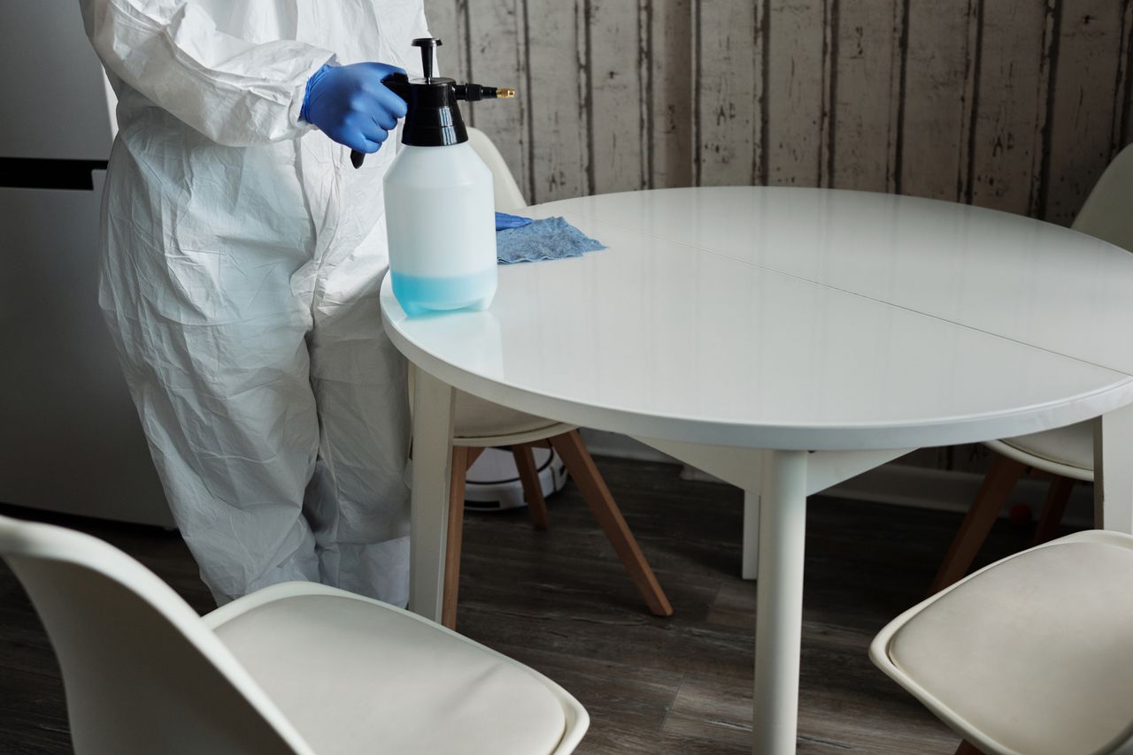 creative-03 Person in protective suit disinfecting a white table indoors with a sprayer.